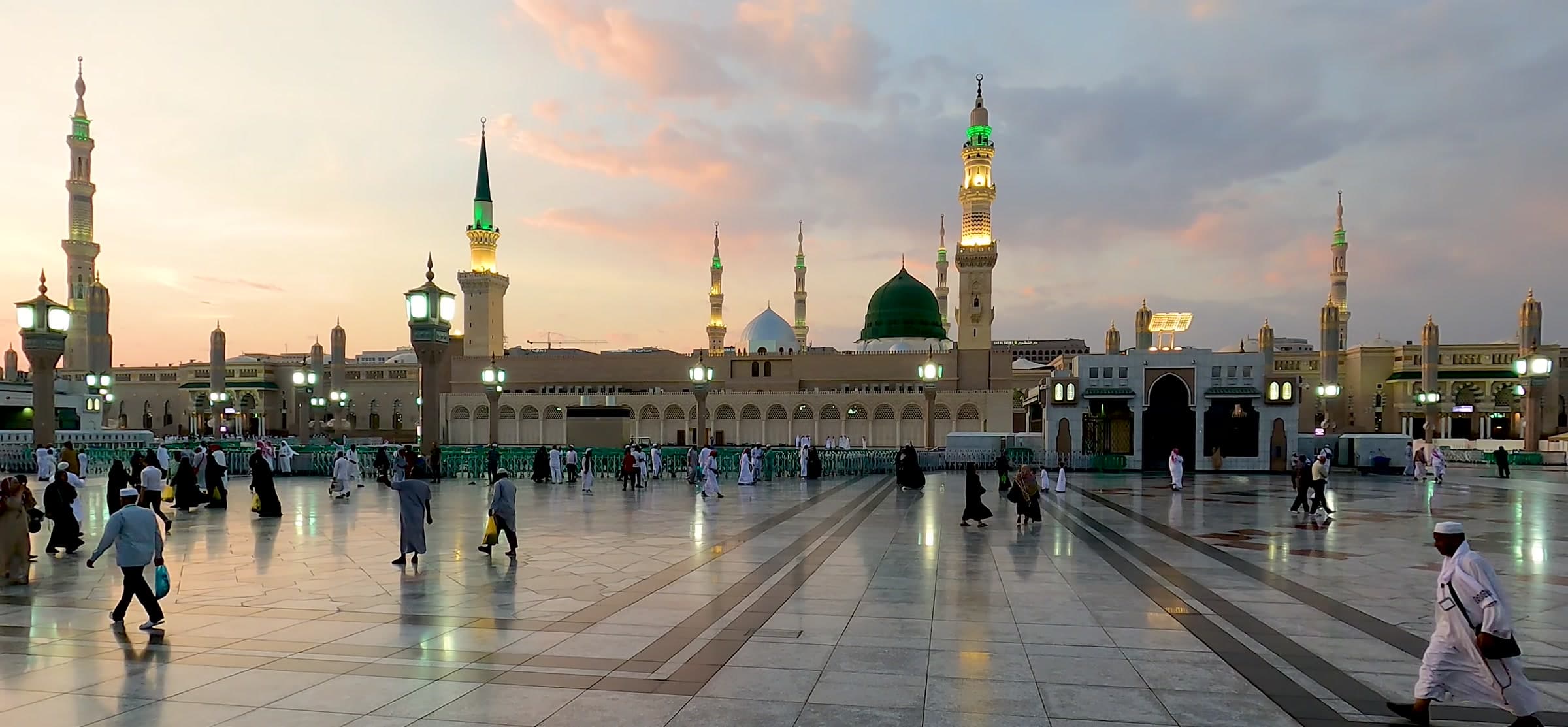 Masjid an-Nabawi at dusk, the green dome and minarets lit against a pink sky.